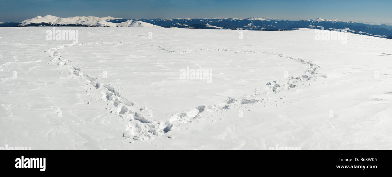 Human footprint form the heart shape on snow covered mountainside plateau and mountain ranges behind. Five shots stitch image. Stock Photo