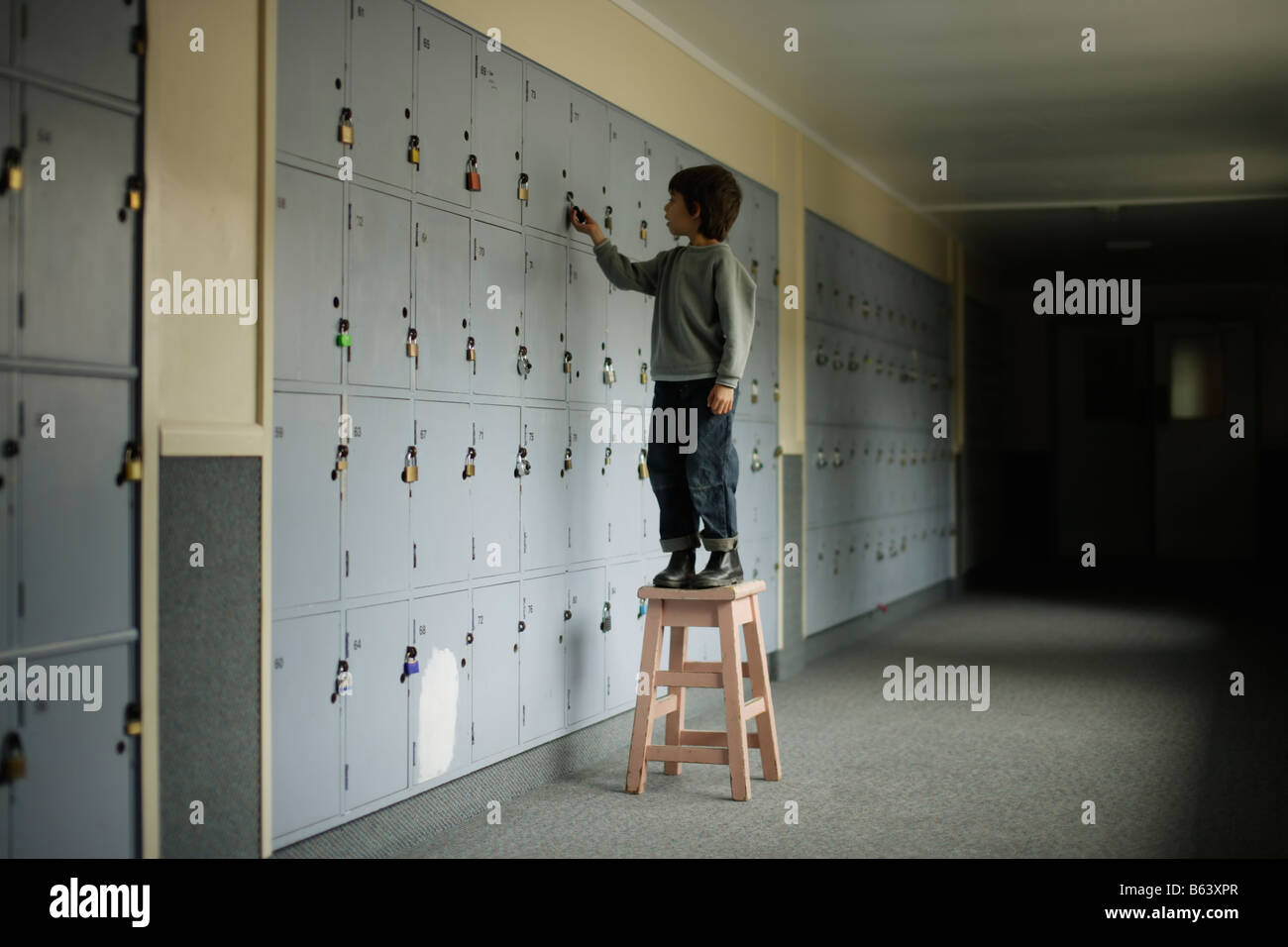 Six year old boy stands on stool in school corridor to reach top locker Stock Photo