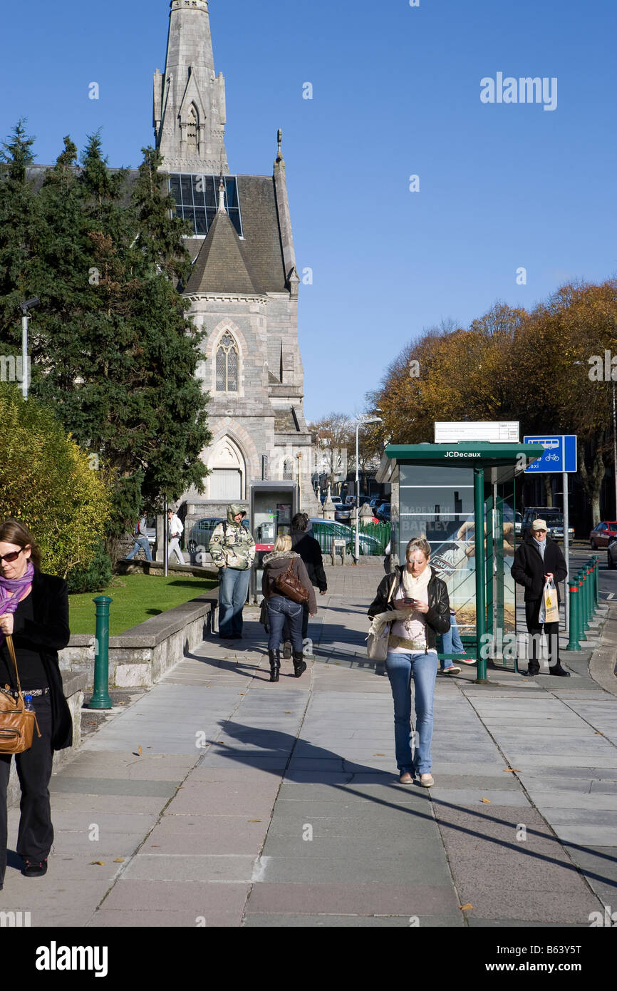 people walking down street. bus stop and church in background in