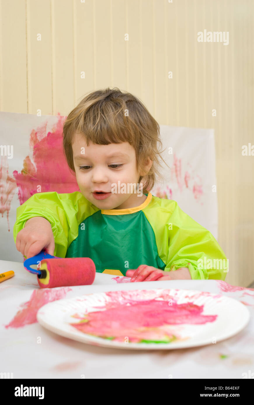 A two year old girl in a plastic smock painting in a playroom with a brush and roller Stock Photo