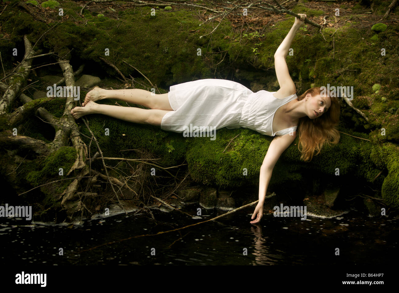 Strawberry blonde woman in diaphanous white cotton slip dress lies adrift at river's edge on a bed of soft green moss. Stock Photo