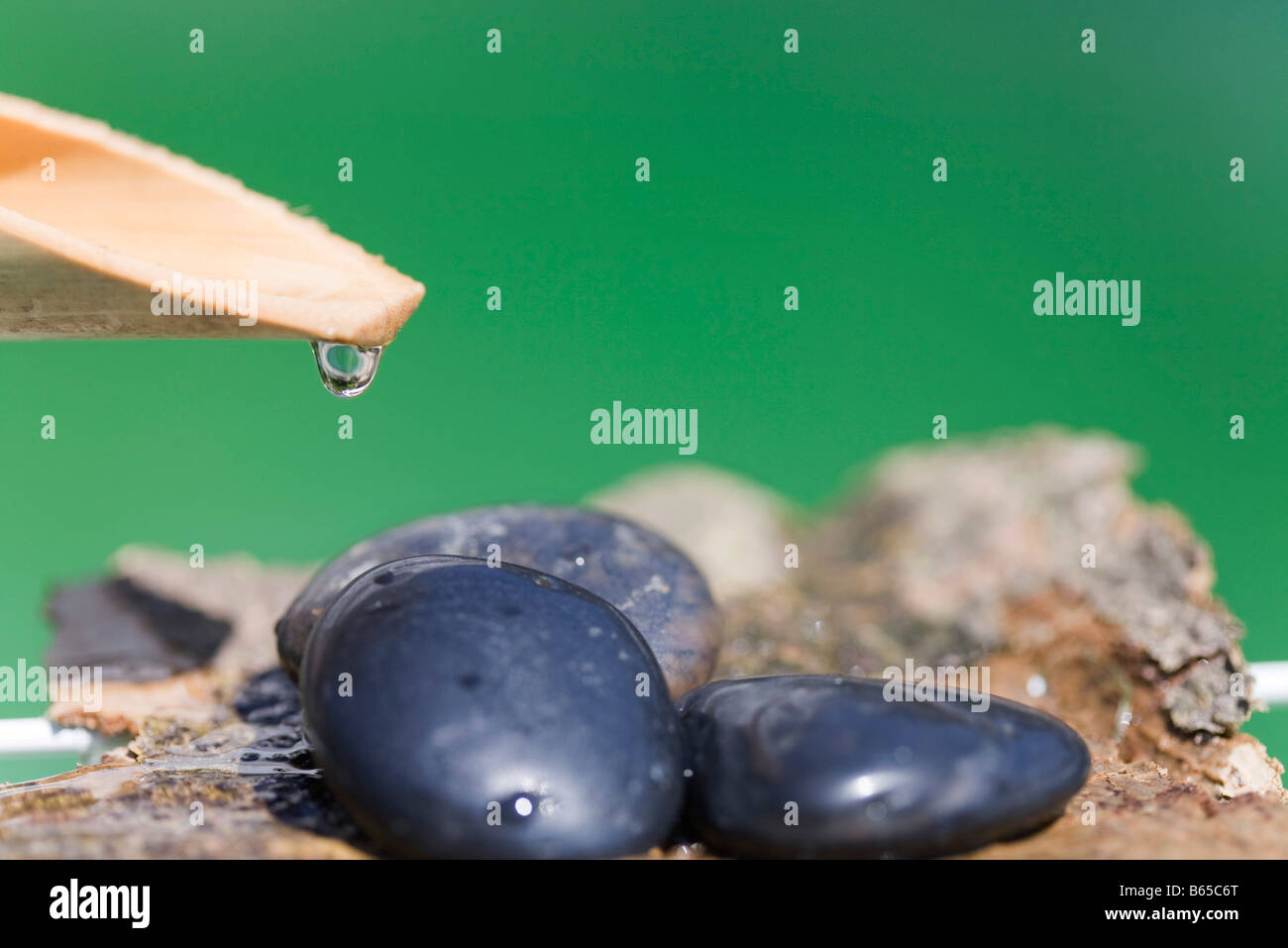 Water droplet hanging off wooden spout over heap of rocks Stock Photo