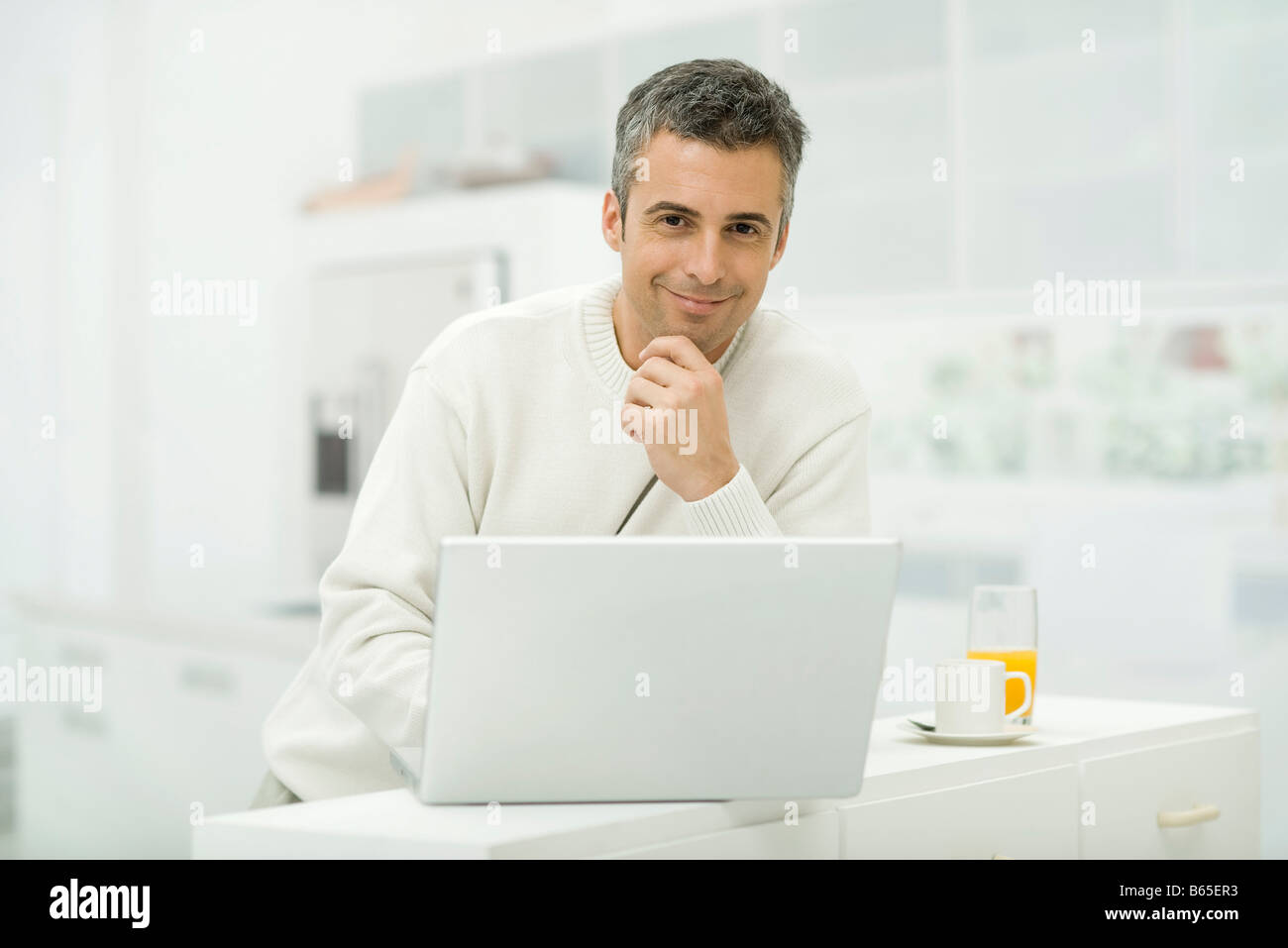 Man using laptop computer in kitchen, smiling at camera Stock Photo