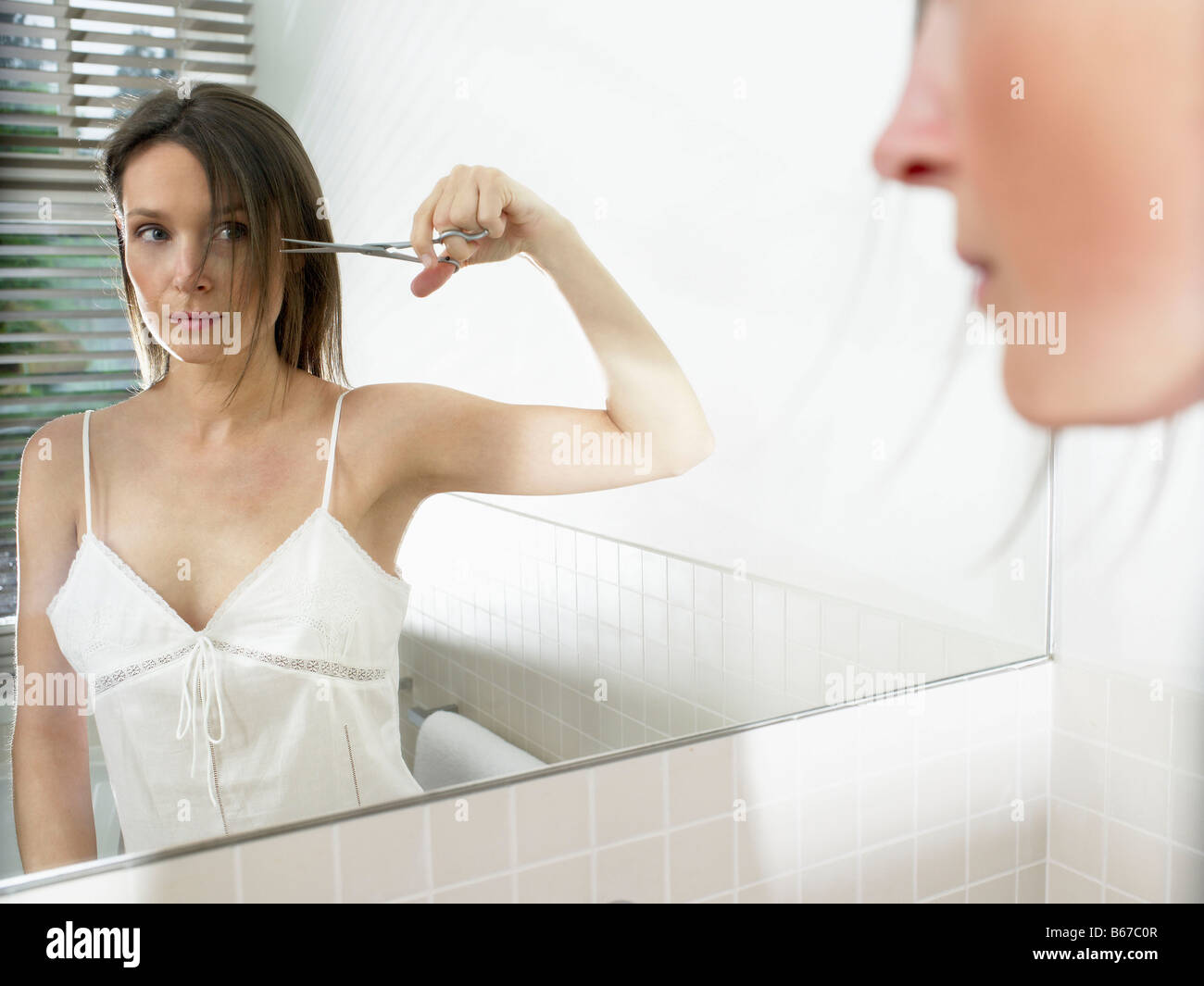 Young woman in home bathroom looking in mirror after cutting her own hair  with scissors Stock Photo - Alamy
