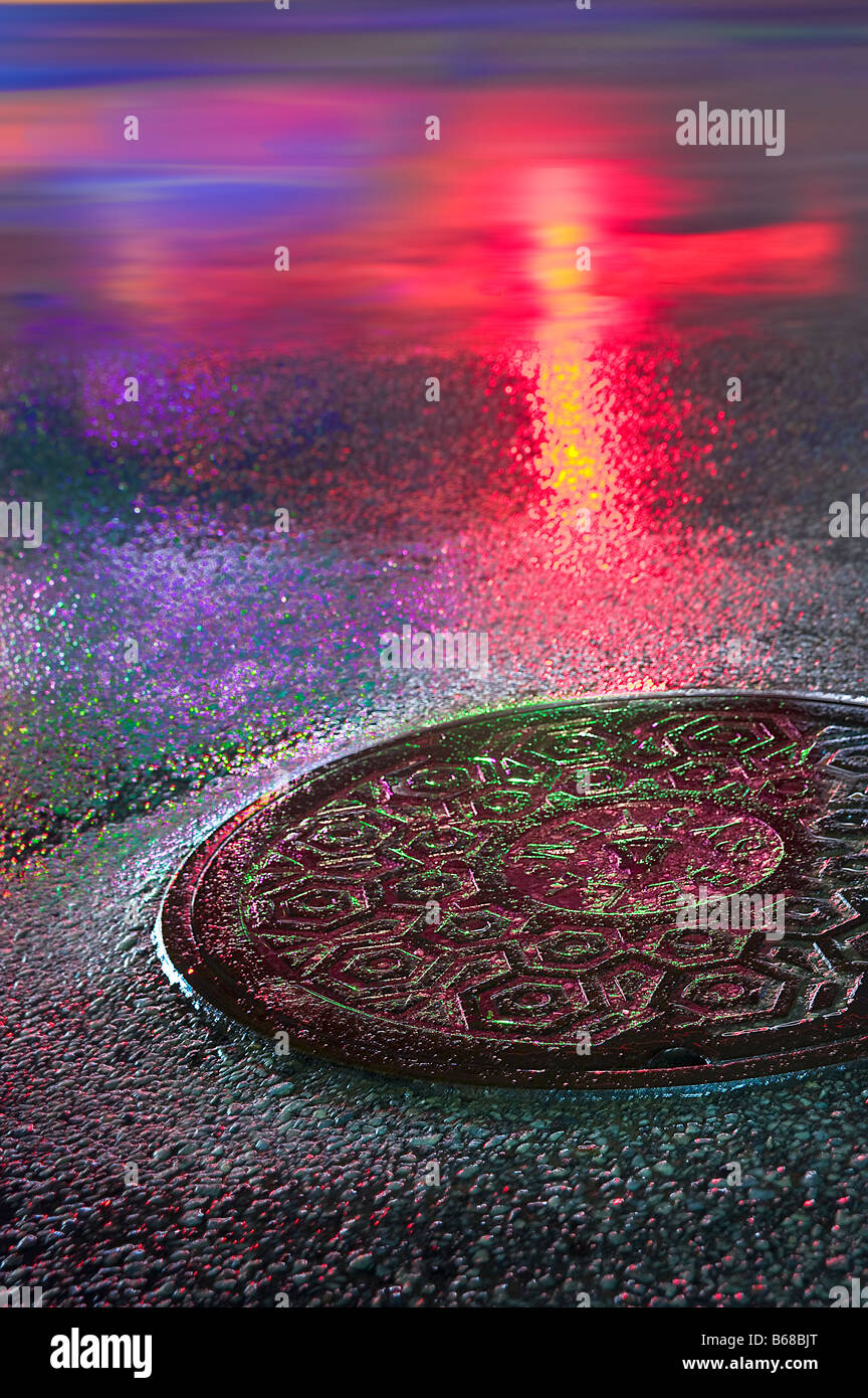 Manhole Cover At Night With Brightly Colored Neon Lights Reflecting Wet Street Pavement Stock Photo