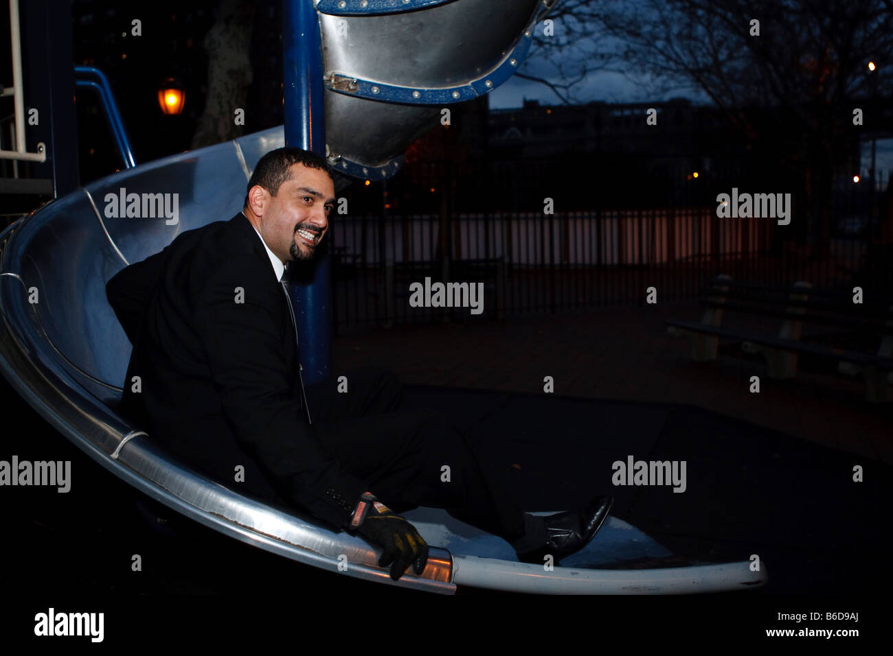 A man in a suit goes down a slide in a children's playground. Stock Photo