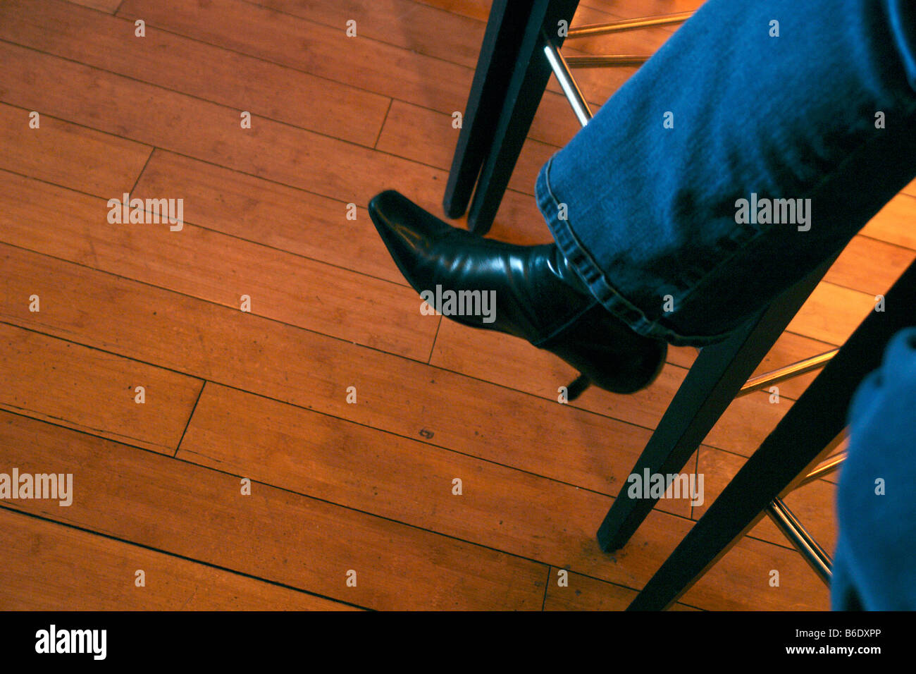 Photograph of the Leg and Foot of a Woman Wearing Blue Jeans and Black Boots Sitting on a Stool on a Wooden Floor Copy Space Stock Photo