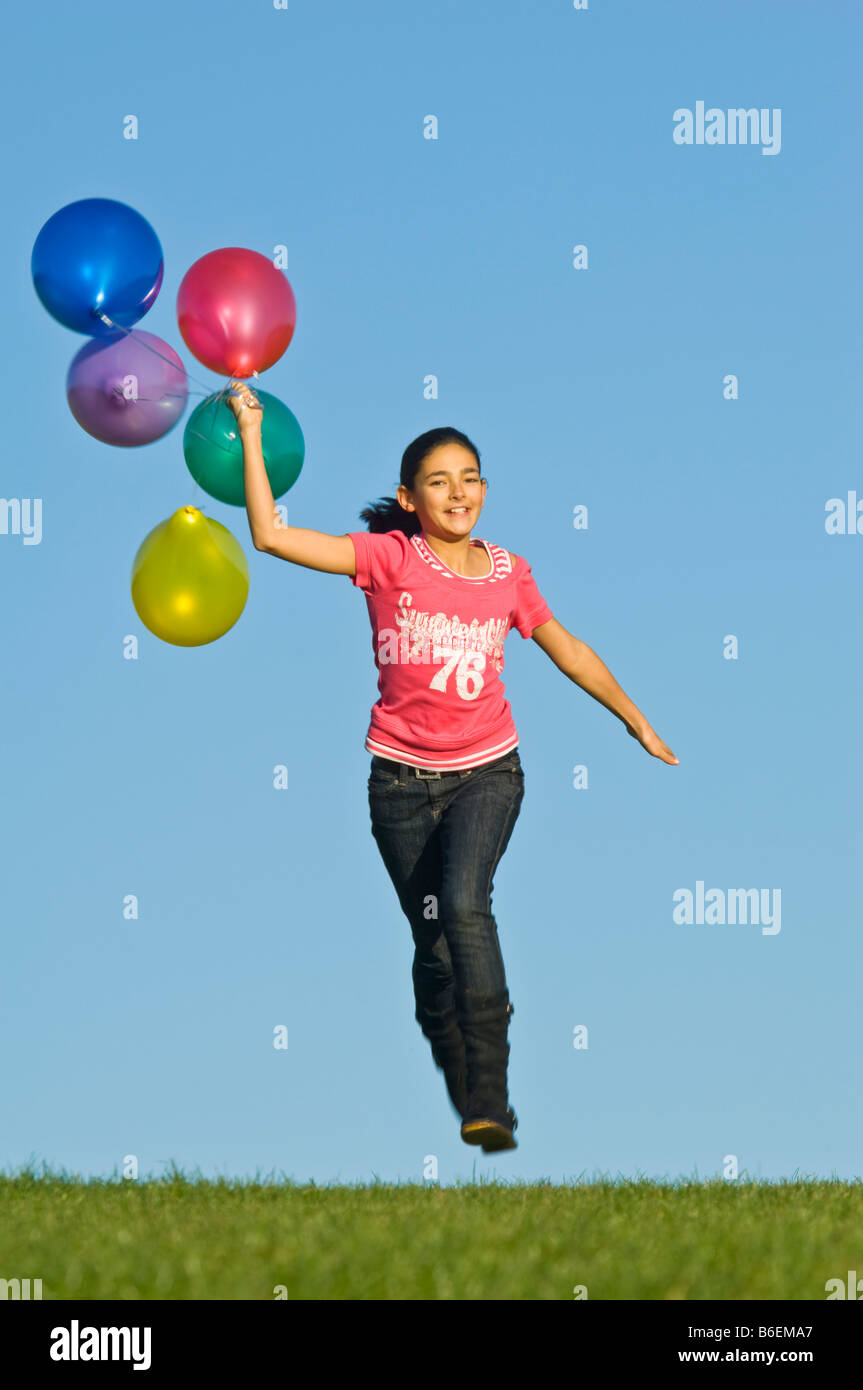 A young girl (11) running happily across green grass with a bunch of colourful balloons trailing behind and blue sky. Stock Photo