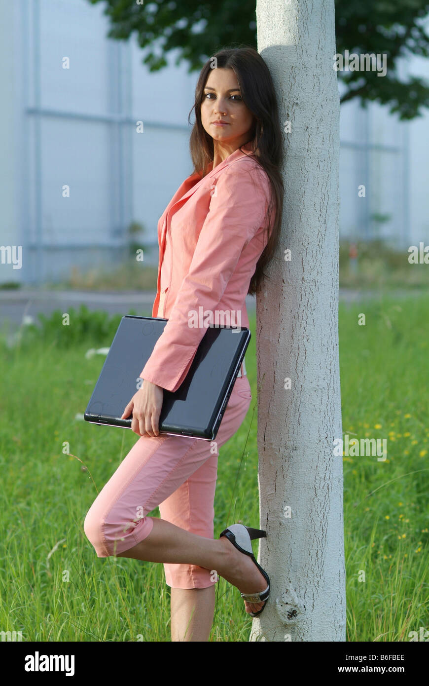 Young woman wearing a pink suit with a laptop under her arm leaning against a white tree trunk Stock Photo