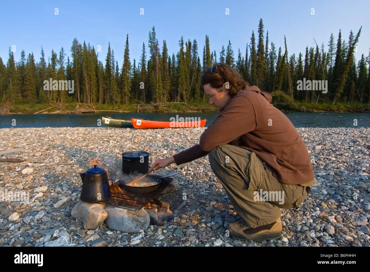 Young man cooking on the camp fire, canoes behind, gravel bar, Liard
