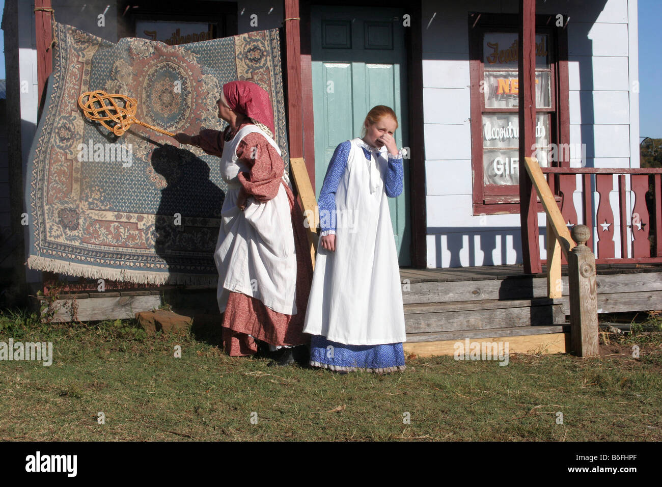 A mother figure is beating the dust from a rug while a young girl is holding her nose Stock Photo