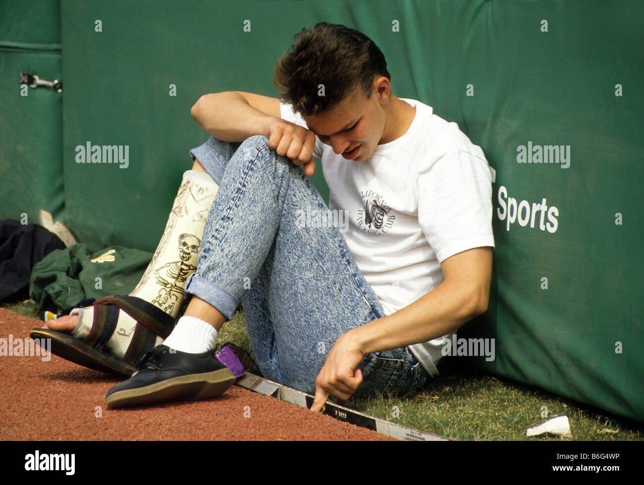 Teen boy with leg in cast sits at edge of sports field unable to play. Stock Photo