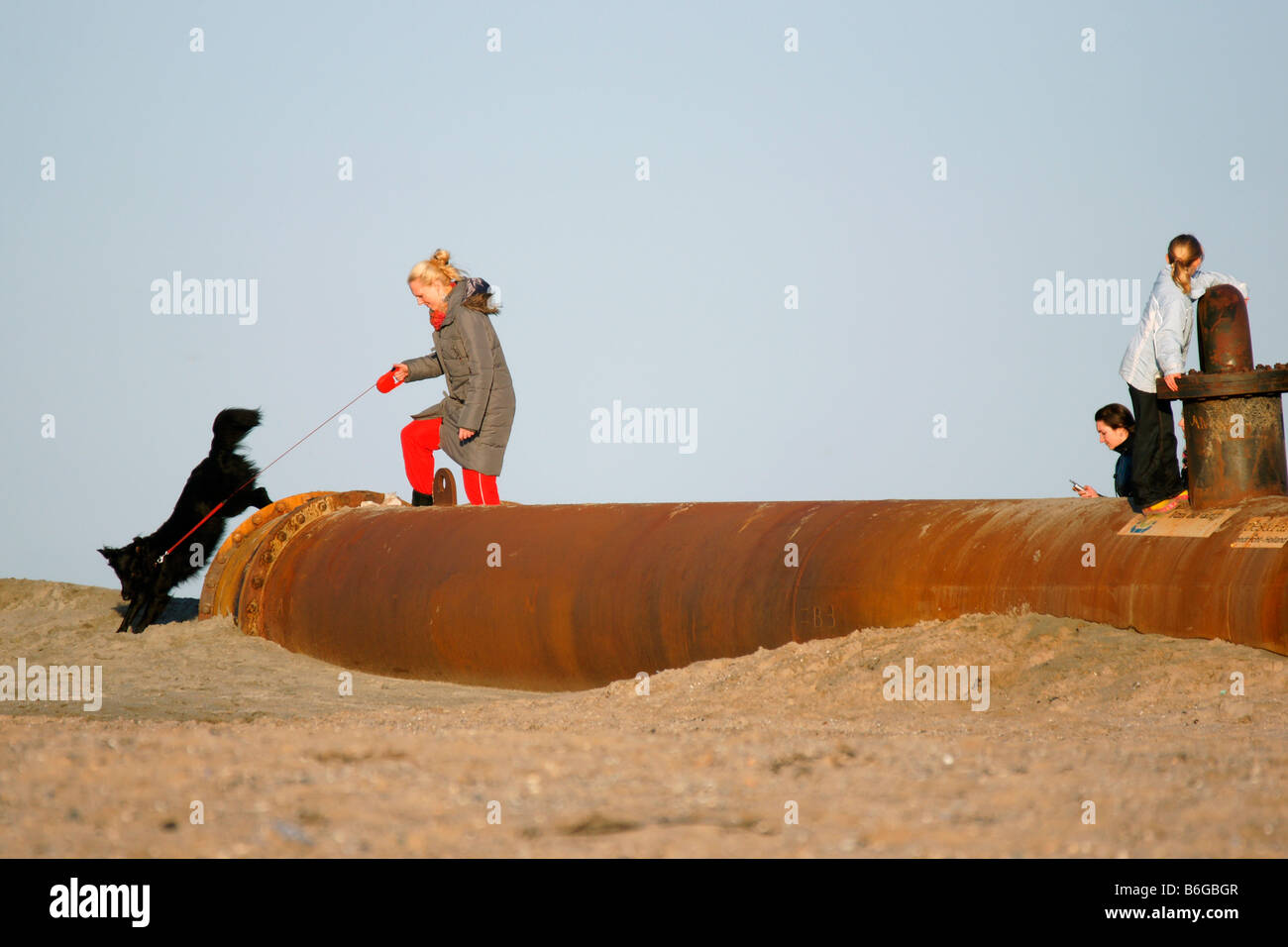 Two children boy girl boys girls walking on huge rusty pipeline with metal valve on sand beach under grey sky in winter Stock Photo
