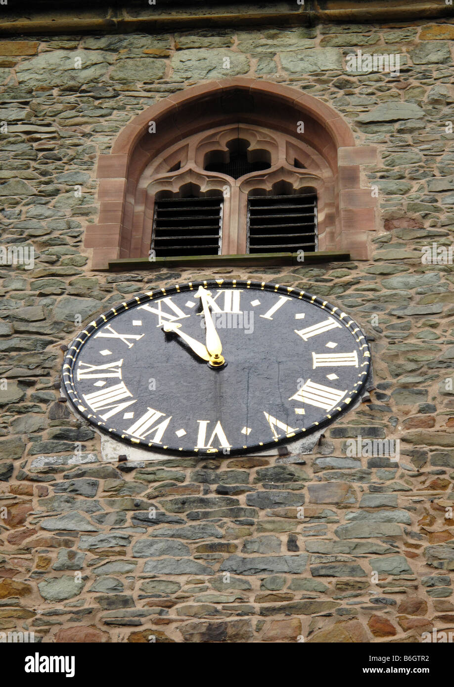 Metal clock face showing 10.59 with roman numerals on stone church clock tower with louvered stone mullion window above. Stock Photo