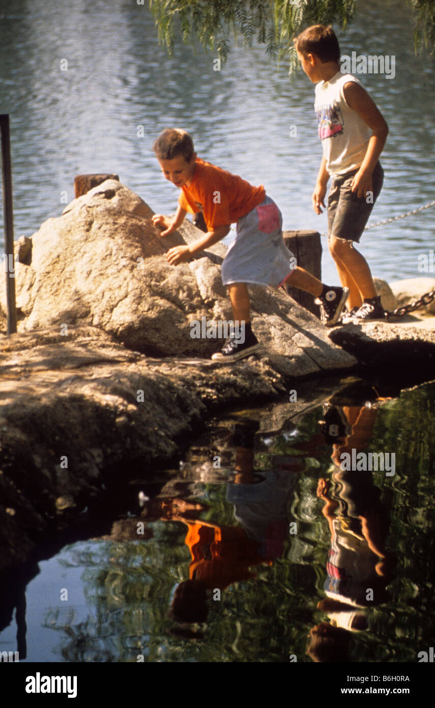 Two young boys carefully walk along edge of water at lake in park. Stock Photo