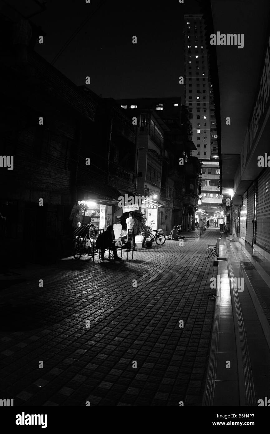 Old side alley in evening in Guangzhou China. Office tower lights reflected on ground, dimly lit neighborhood shop. Stock Photo