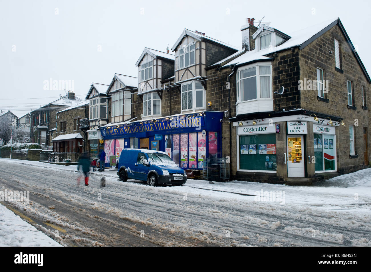 The local village shops on high street after a fall of snow - people walking on snowy pavement & road - Burley in Wharfedale, Yorkshire, England, UK. Stock Photo
