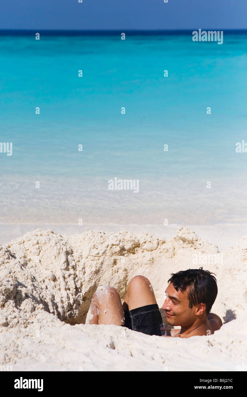 A young man in sitting in a shallow hole on a beach in The Maldives Stock Photo