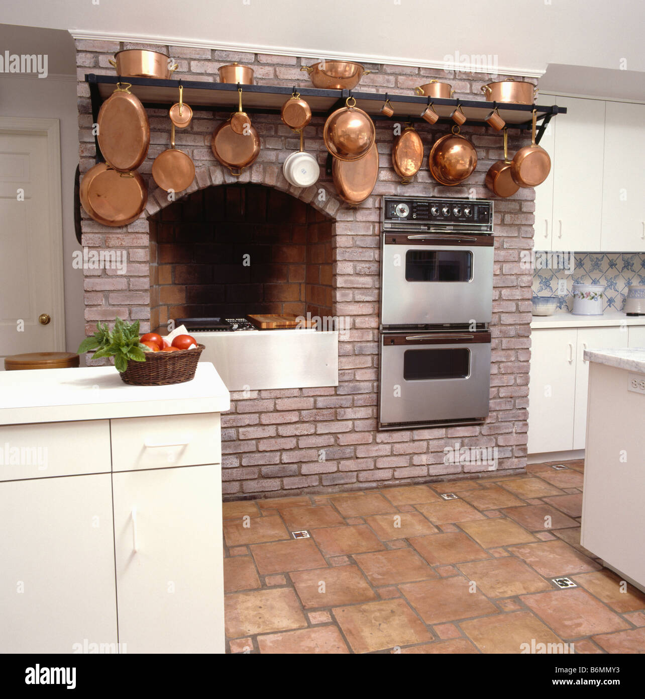 Copper pans on rack above double ovens and hob fitted into brick fireplace alcove in kitchen with terracotta floor tiles Stock Photo