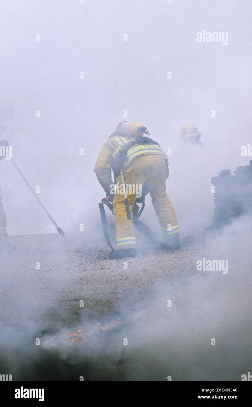 Fireman wearing full protective gear cuts holes in roof of burning house to vent smoke. Stock Photo