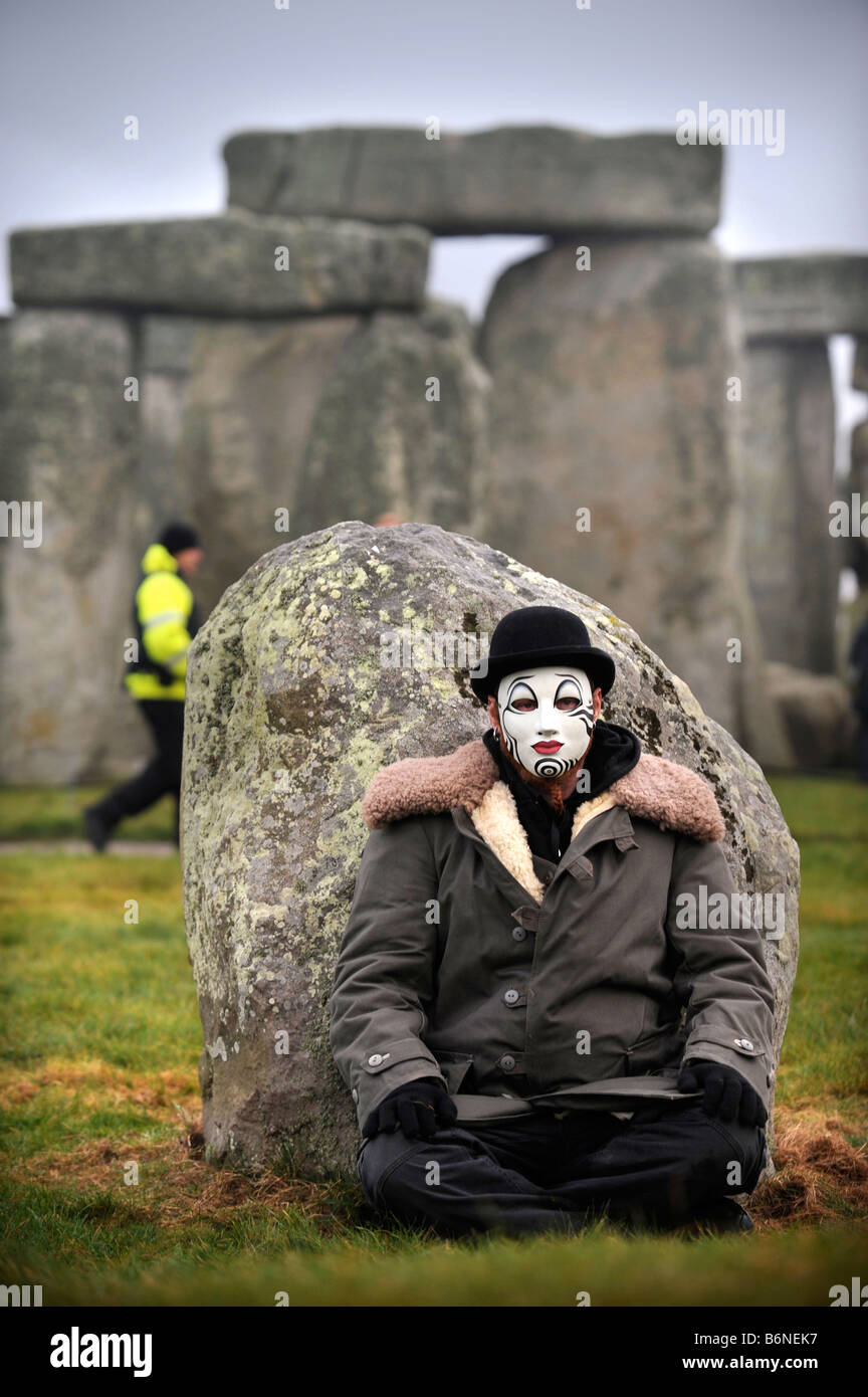 A VISITOR TO STONEHENGE WEARING A WHITEFACE CLOWN MASK SITS BY AN OUTER STATION STONE WHILE A SECURITY GUARD APPEARS TO BE SEARC Stock Photo