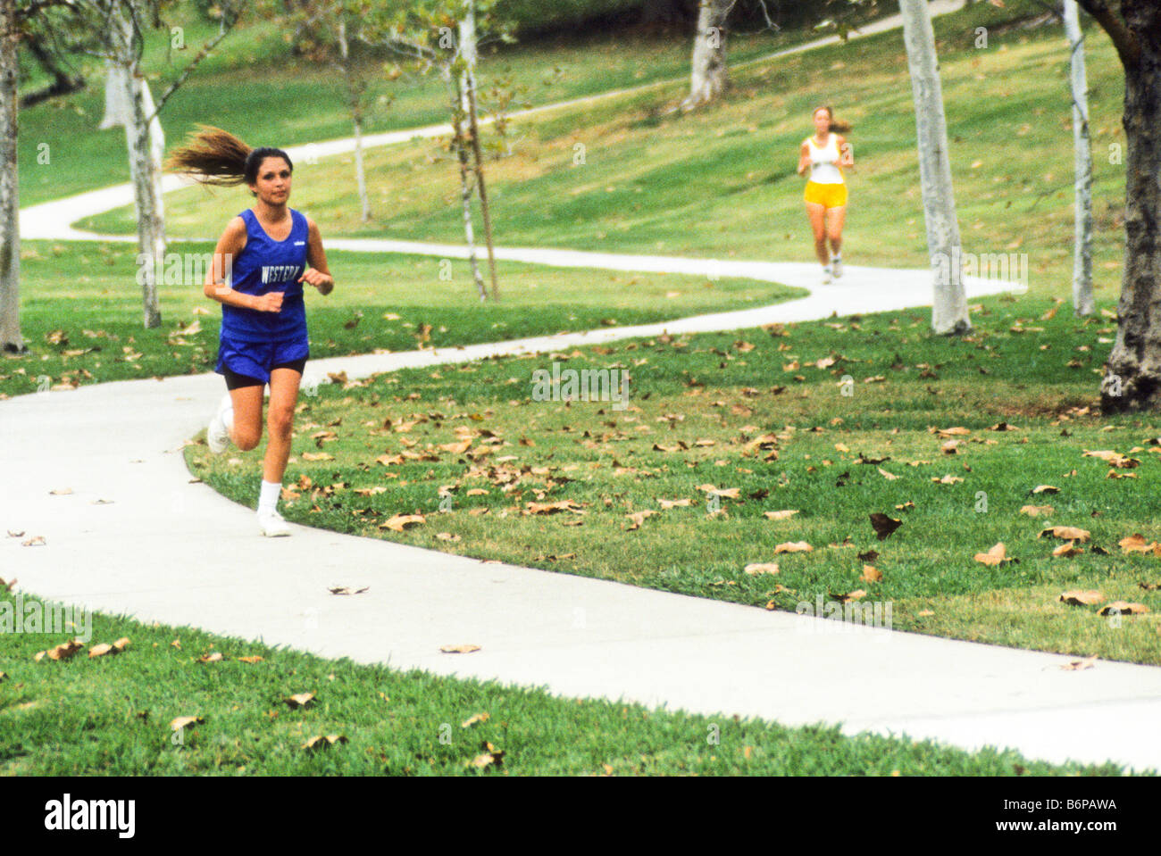 Two high school girls run down curved sidewalk in park during cross country race. Stock Photo