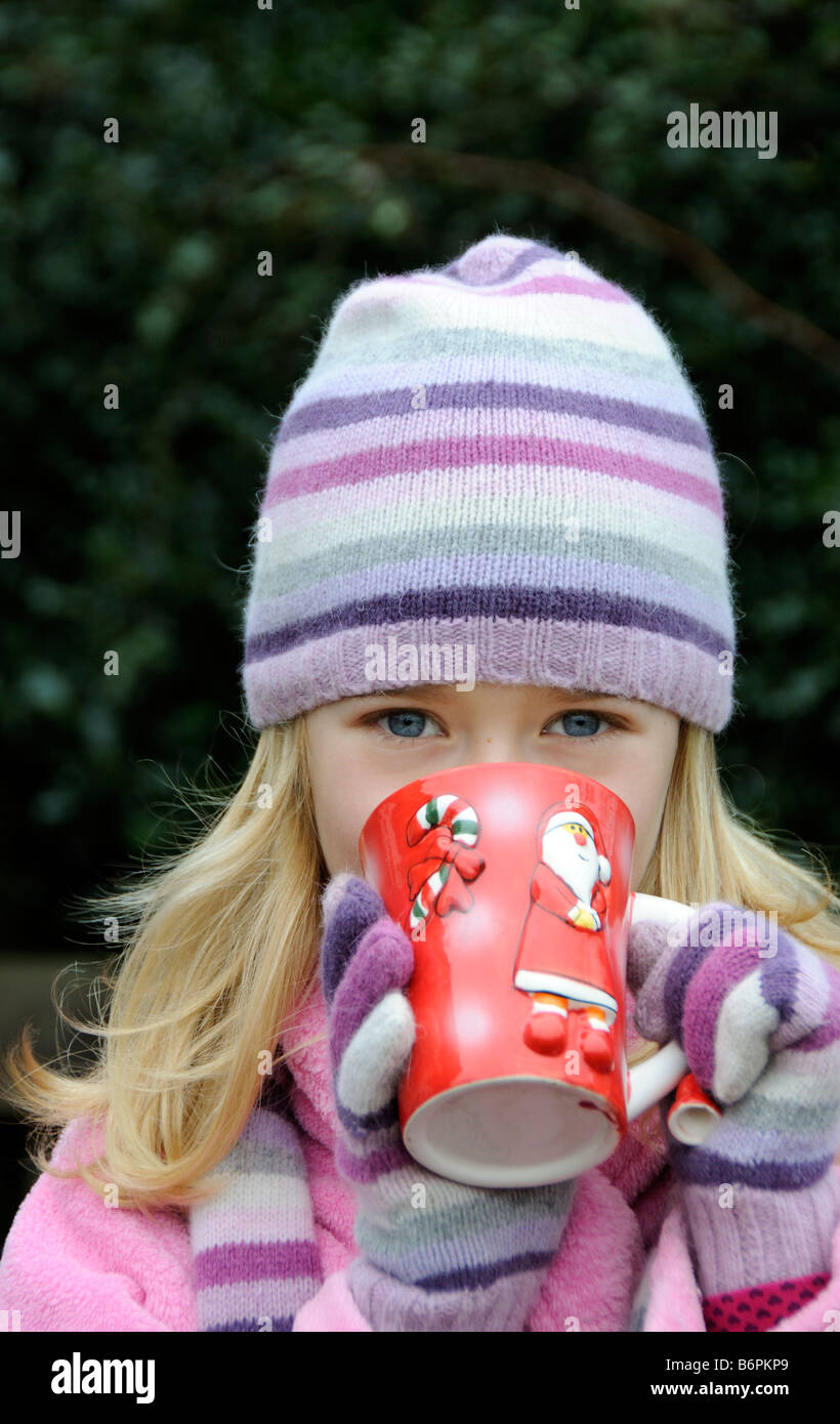 Portrait of a little girl smiling and wearing warm clothes with a woolly hat and drinking from a mug Stock Photo