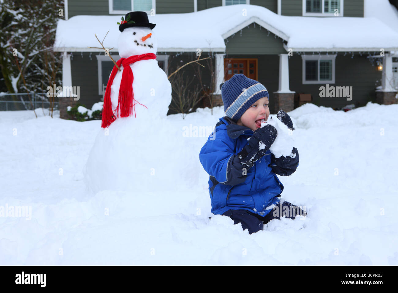 Young boy eating snow with snowman and home in background Stock Photo
