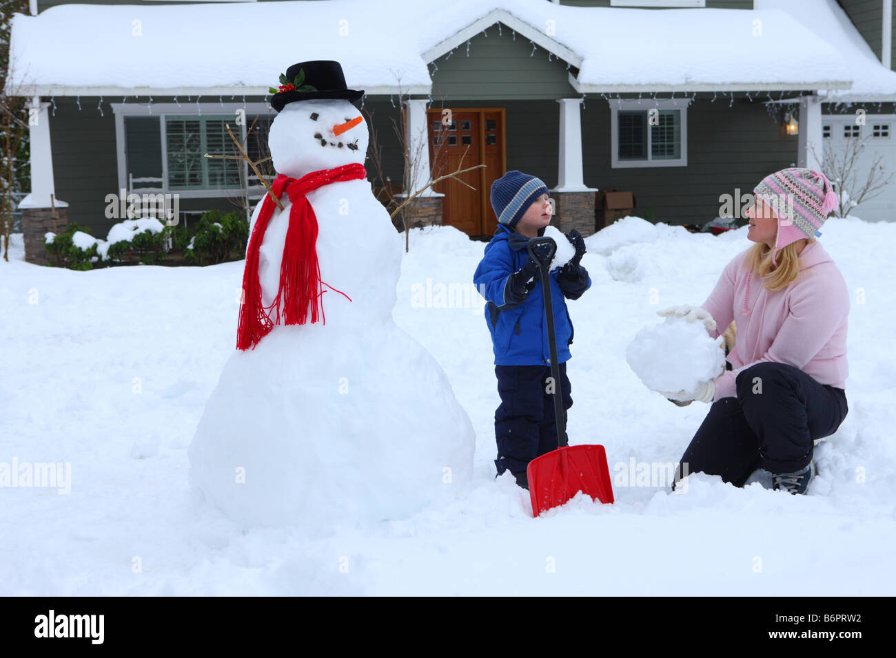 Mother and young boy playing in snow by snowman and house Stock Photo