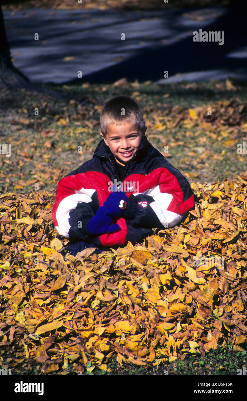 Young boy wearing heavy jacket sits in pile of autumn leaves in his yard. Stock Photo