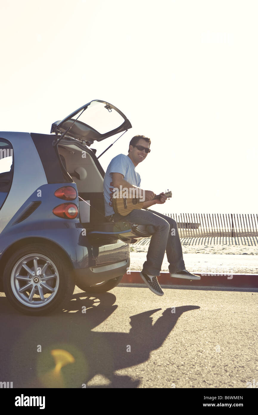Young man seated on boot of Smartcar at beach playing guitar Stock Photo
