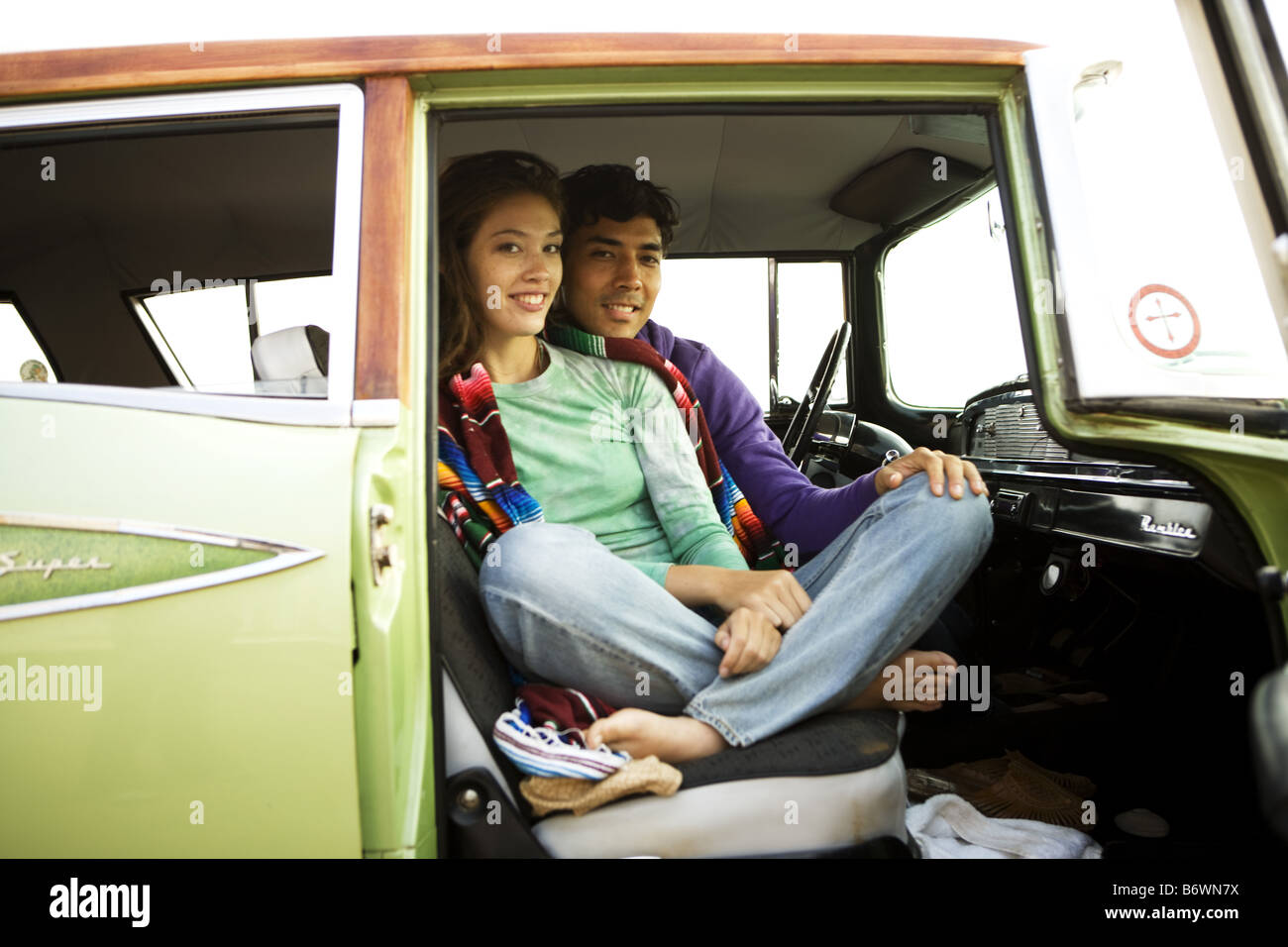 smiling surfer couple sit in vintage car on cliff at beach Stock Photo