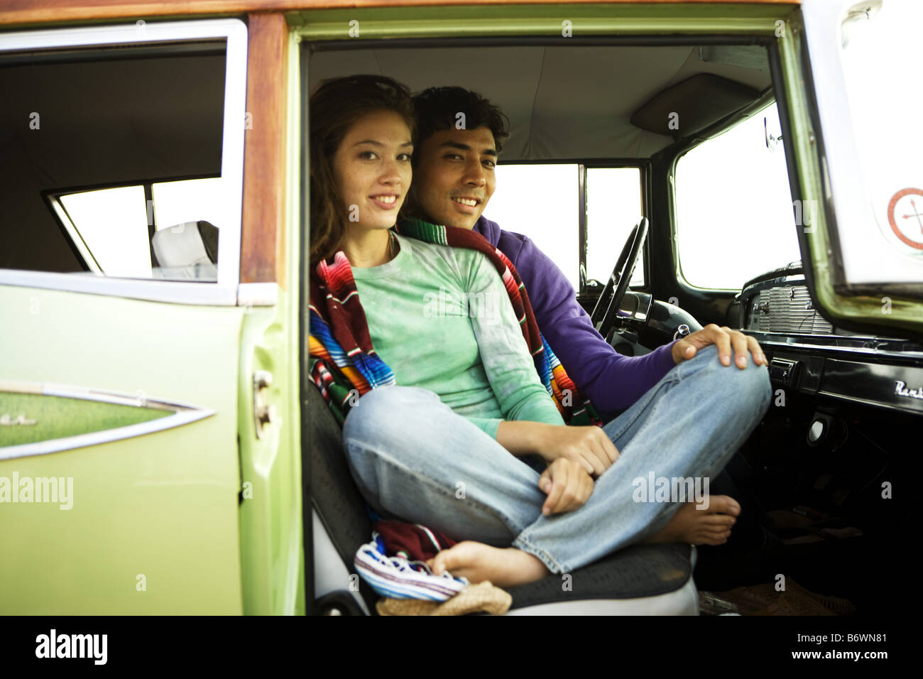 smiling surfer couple sit in vintage car on cliff at beach Stock Photo