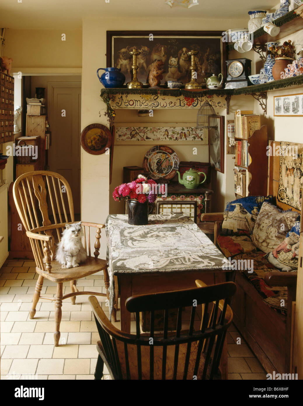 Cat on antique Windsor chair in cluttered cottage dining room with tapestry cushions on wooden settle and crockery on shelves Stock Photo