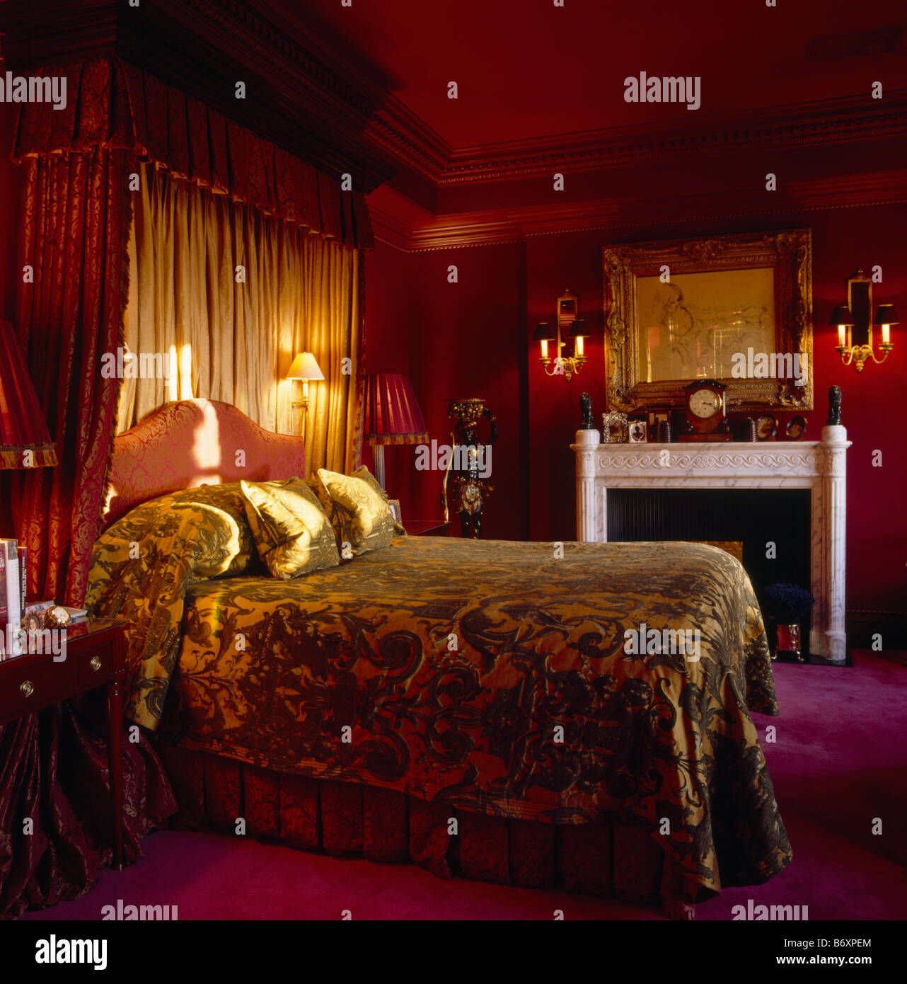 Velvet bedcover on half tester bed with lighted wall lights and cream drapes in dark red bedroom with marble fireplace Stock Photo