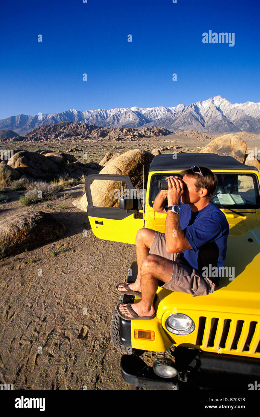 Man sitting on a jeep in the desert next to mountains with binoculars