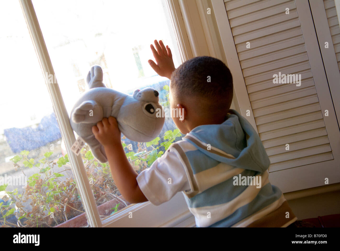 A young boy holding a teddy bear looking out of a window Stock Photo