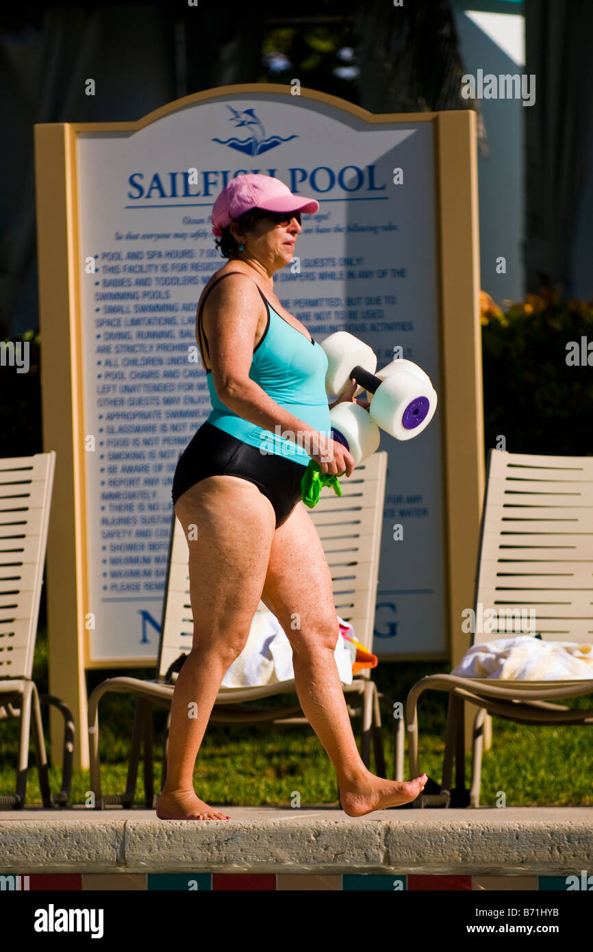 Palm Beach Shores , middle aged large lady in green swimming costume & pink baseball cap walks by pool with exercise weights USA Stock Photo