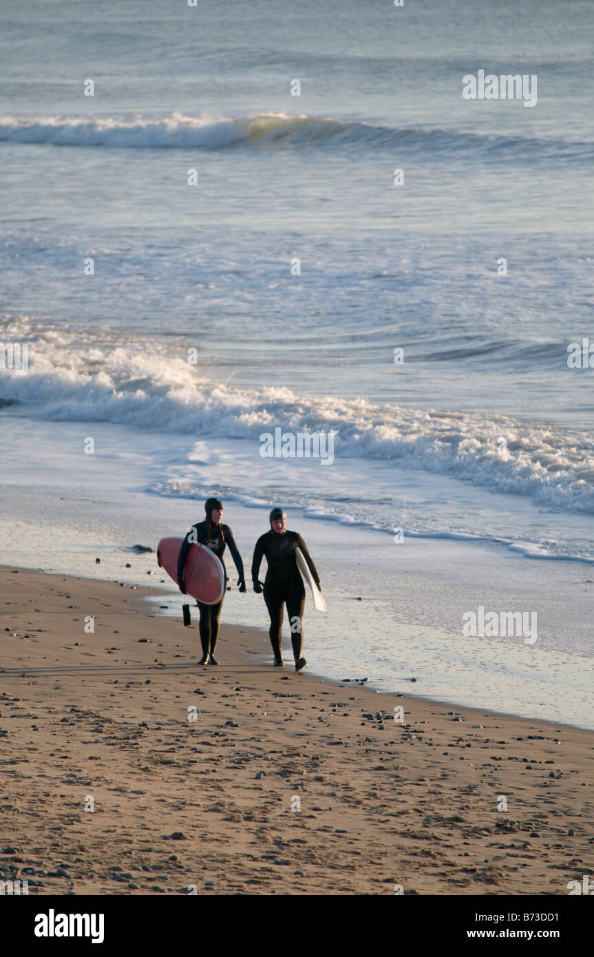 Two young women wearing wet suits and carrying surf boards Tywyn Gwynedd Cardigan Bay wales UK, January afternoon Stock Photo