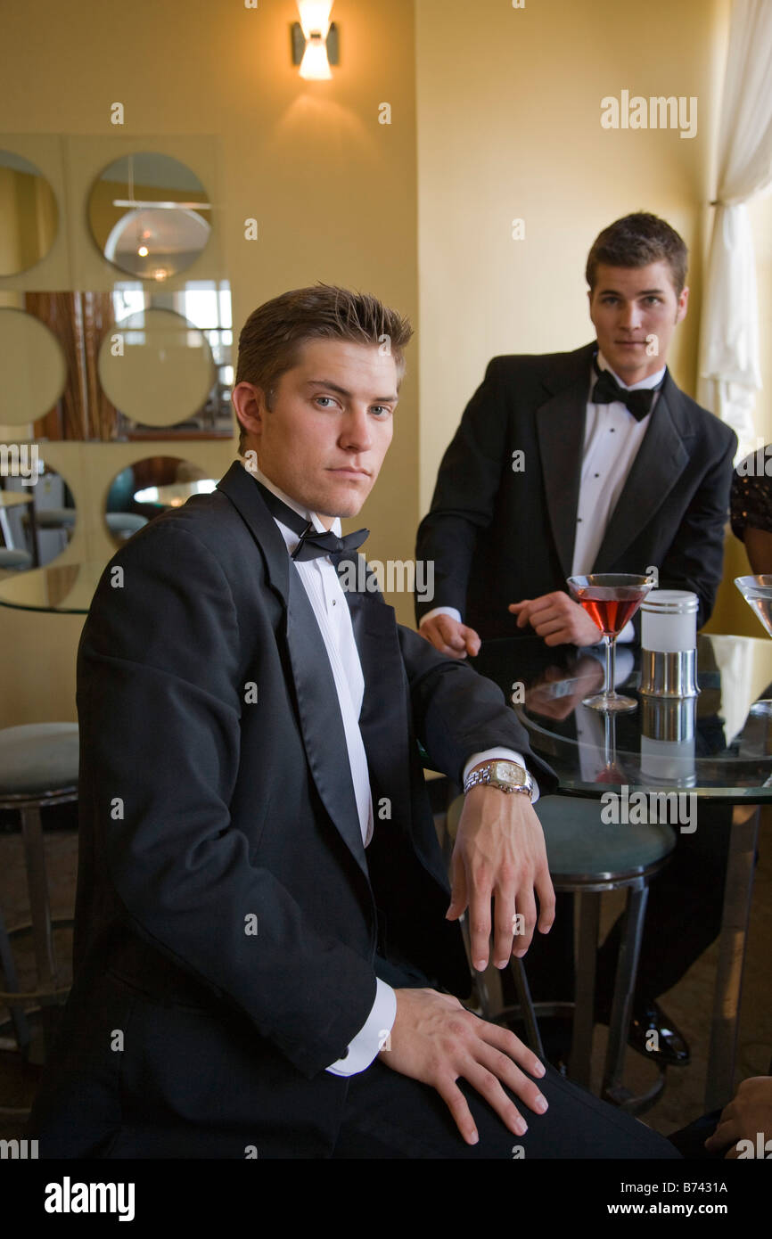 Two young adult men wearing tuxedos having drinks in restaurant lounge