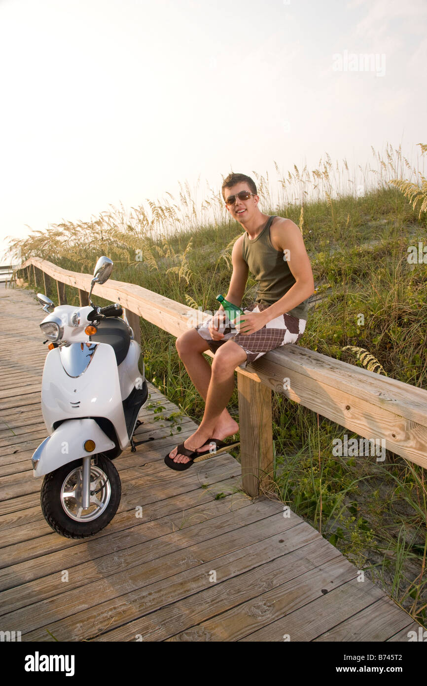Young man sitting on railing of wooden boardwalk by parked moped Stock Photo