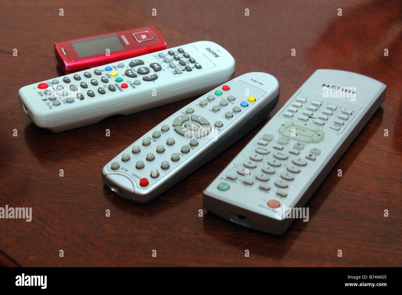 A red mobile phone lies on a table alongside remote controls for digital T.V, DVD. Stock Photo