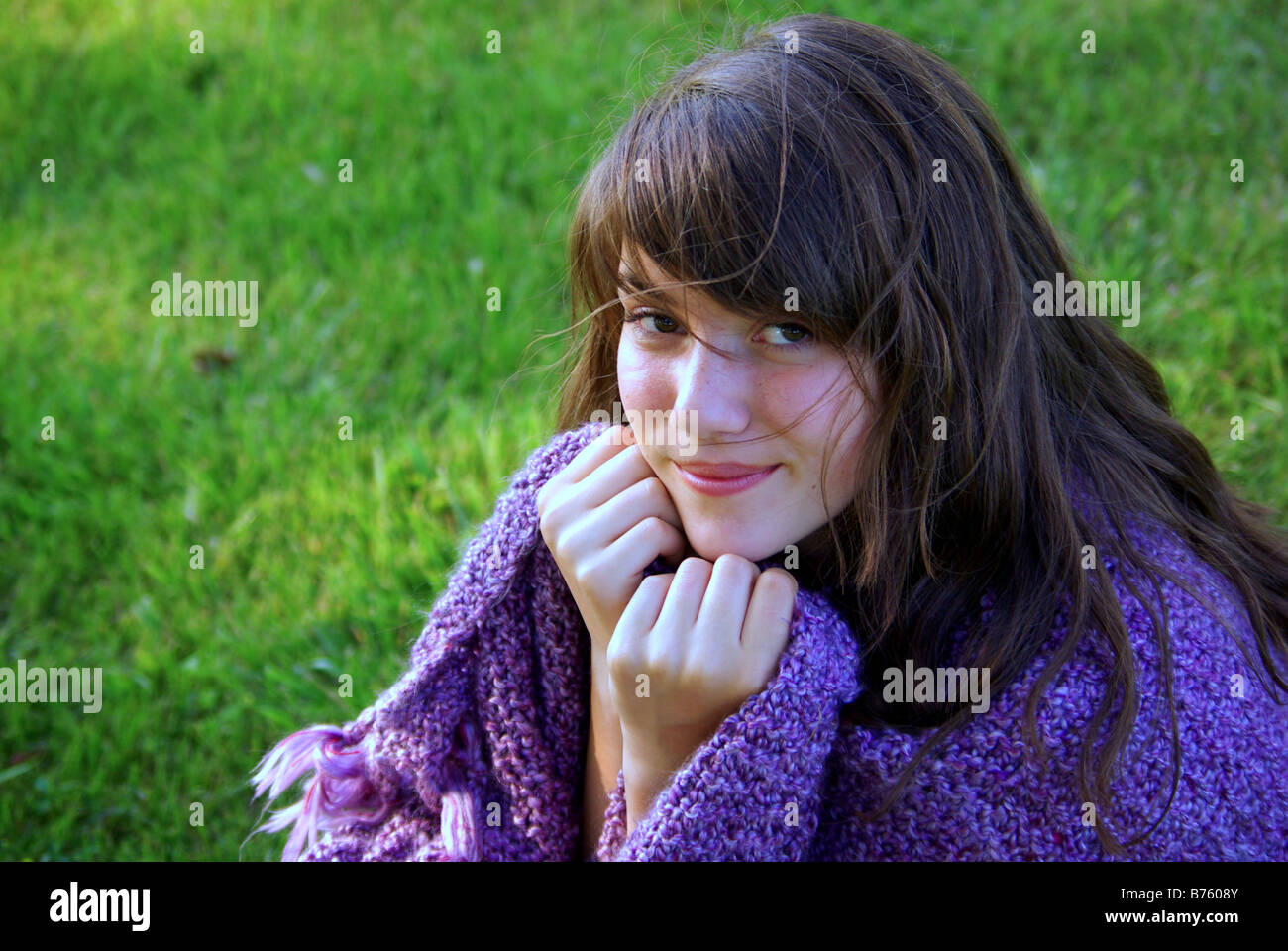 A beautiful fourteen year old girl sits on the grass warpped in a purple handknit shawl. Stock Photo