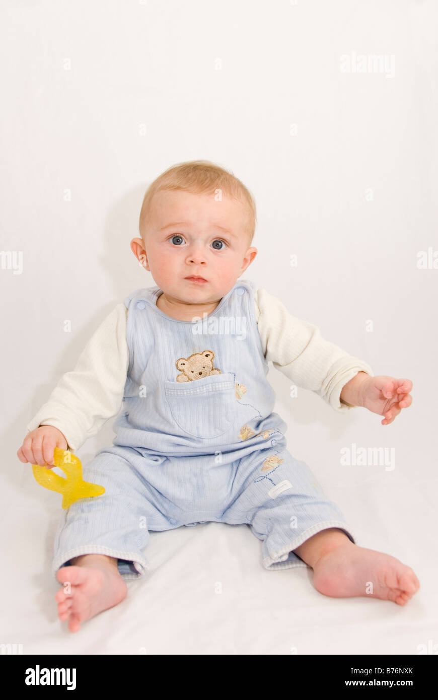 Full Length Portrait of Worried Frowning Baby Boy Sitting Up Holding Yellow Teething Toy Cut Out on White Stock Photo