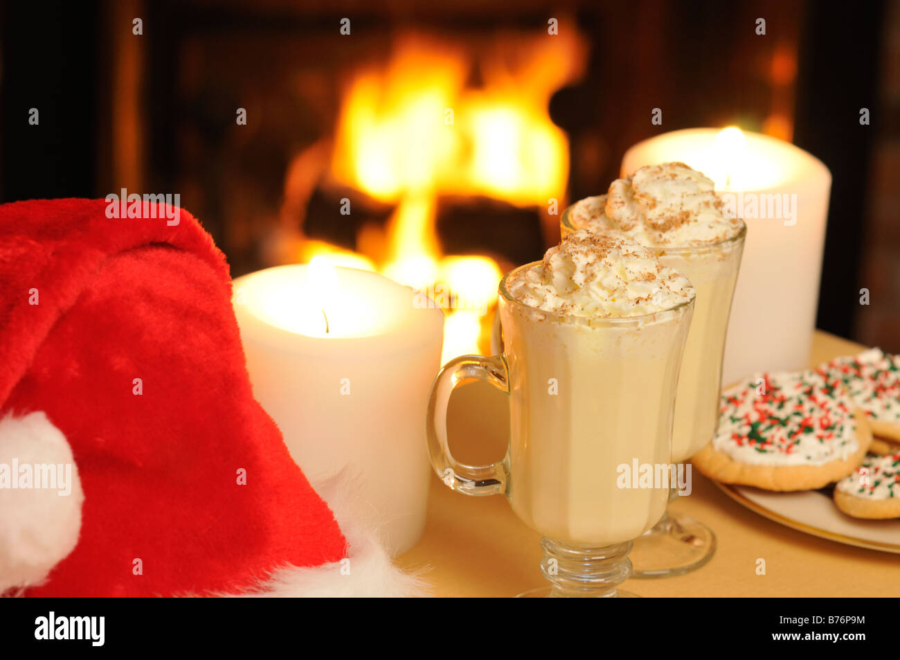 Glass mugs of Christmas eggnog with decorated cookies,  candles and santa hat with fireplace burning in background Stock Photo