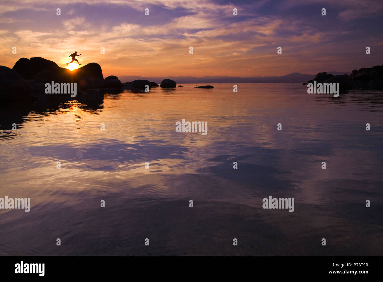 A man jumping on a rock on the shore of Lake Tahoe in California at sunset with reflecting water Stock Photo