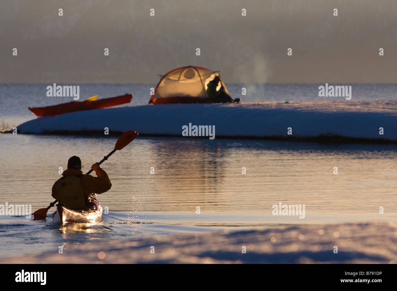 A man sea kayaking past a woman camping on the snowy shore of Lake Tahoe in California in winter. Stock Photo