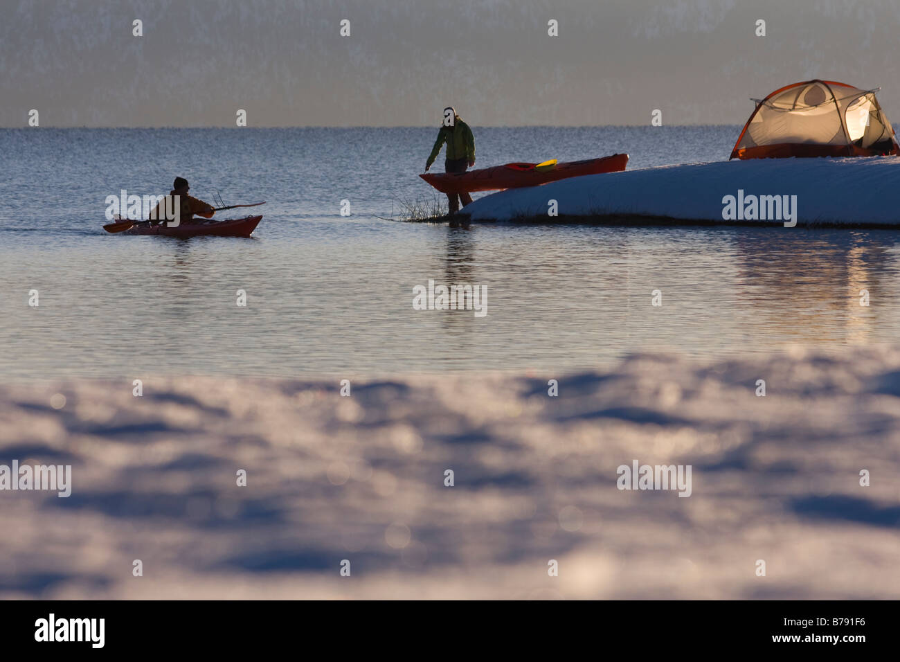 A man in a sea kayak paddling along the shore near a woman and a tent on Lake Tahoe in California on a snowy day at sunrise Stock Photo