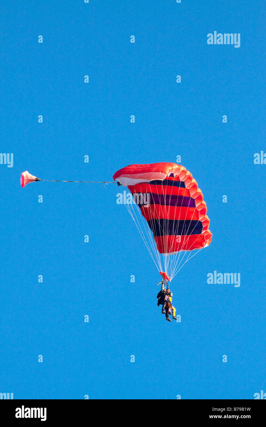 TANDEM PARACHUTING IN ENGLAND A RED STRIPED PARACHUTE CARRYING TWO PEOPLE GLIDES ACROSS THE BLUE SKY Stock Photo
