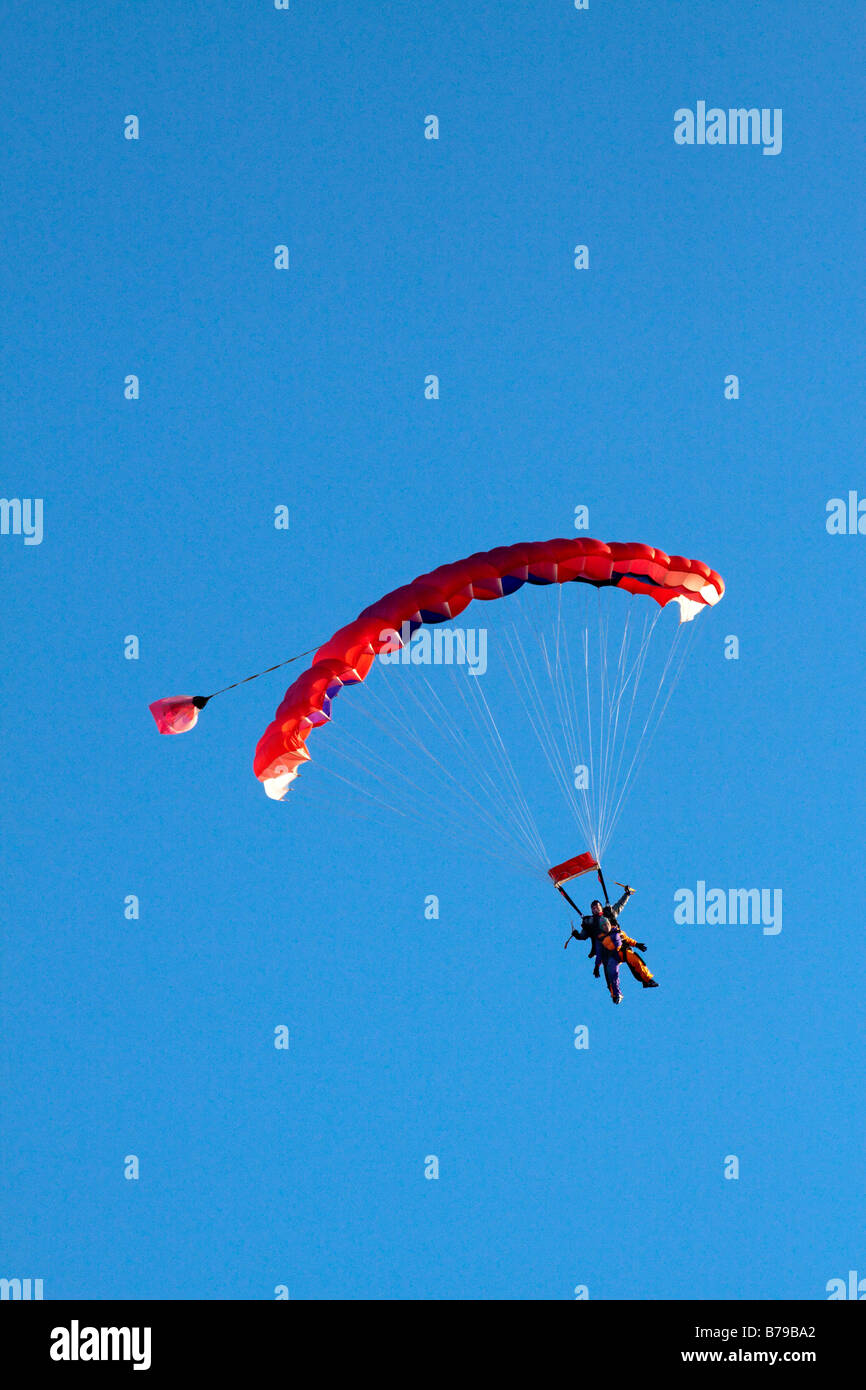 TANDEM PARACHUTING IN ENGLAND A RED STRIPED PARACHUTE CARRYING TWO PEOPLE GLIDES ACROSS THE BLUE SKY Stock Photo